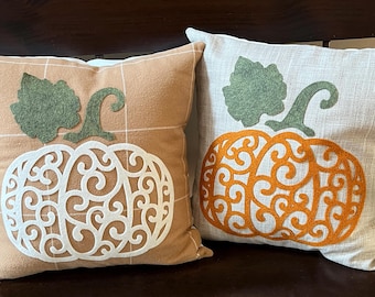 May include: Two decorative throw pillows with a pumpkin design. The pillows are made of a beige and white fabric with a brown plaid pattern. The pumpkins are made of felt and have a white and orange lace design.