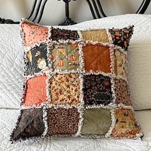 May include: A square rag quilt pillow featuring a patchwork design of various autumn-themed fabrics. The squares display patterns of pumpkins, scarecrows, fall leaves, and solid orange and brown textures, all joined with frayed seams.