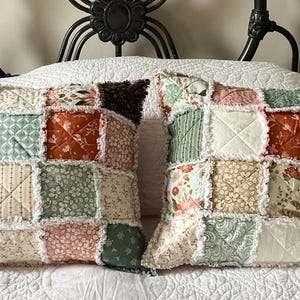 May include: Two square throw pillows featuring a patchwork design with frayed seams. The fabric squares display various floral and geometric patterns in shades of sage green, burnt orange, cream, and brown, with visible diagonal quilting stitches.