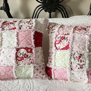 May include: Two square patchwork pillows with a ruffled edge. The pillows feature a mix of pink, red, and white fabrics with floral and bird patterns. The pillows are placed on a white quilted bedspread.