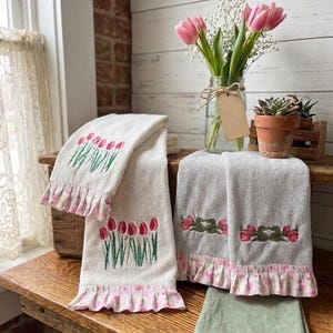 May include: Cream-colored embroidered tulip towels with pink ruffled edges. The towels feature red tulip embroidery with green stems and leaves. A green folded washcloth is also present.