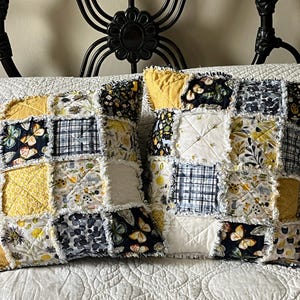 May include: Two square rag-style throw pillows featuring a patchwork design of quilted fabric squares in shades of mustard yellow, navy blue, white, and grey. The patterns include butterflies, floral motifs, and plaid, with frayed seams between each square.