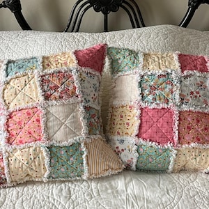 May include: Two decorative patchwork pillows with a variety of floral and geometric patterns in shades of pink, blue, and cream. The pillows have a ruffled edge and are placed on a white bedspread.