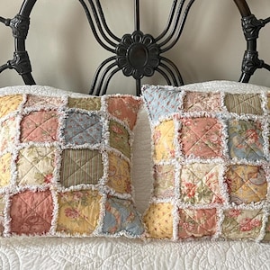 May include: Two square throw pillows with a patchwork design. Each pillow features a variety of pastel-colored floral and patterned fabrics, including pink, yellow, and blue. The edges are finished with a frayed, white trim, creating a rustic, vintage aesthetic.
