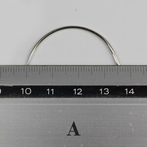 May include: A close-up of a curved, silver needle measuring approximately 12 inches long. The needle is placed on a ruler with markings from 10 to 14 inches.