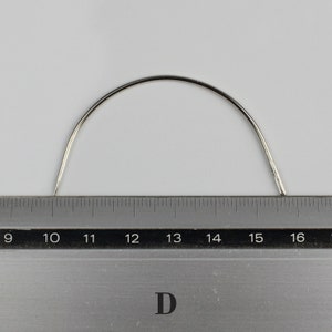 May include: A silver curved needle measuring 16 inches in length, placed on a ruler.