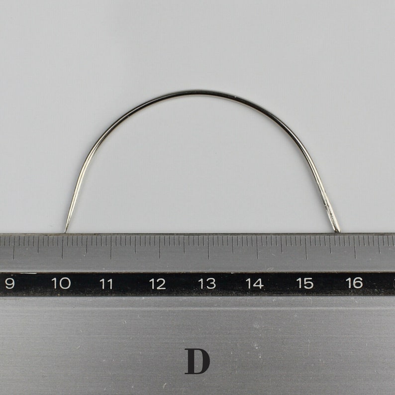 May include: A silver curved needle measuring 16 inches in length, placed on a ruler.