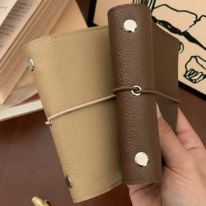 Könnte beinhalten: Zwei kleine Notizbücher mit lederartiger Textur, eines in Beige und eines in Dunkelbraun. Jedes Notizbuch verfügt über einen vertikalen Gummibandverschluss und zwei silberfarbene Metallnieten am Buchrücken. Die Einbände haben eine genarbte Oberfläche.