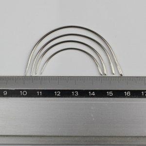 May include: Four silver curved needles of different sizes, measured against a ruler with markings from 9 to 17 inches.