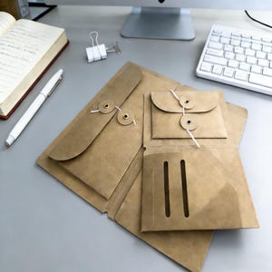 Könnte beinhalten: Ein Dokumentenorganizer aus hellbraunem Papier mit einer großen Klappe mit Schnur- und Knopfverschluss, einem kleineren angebrachten Umschlag mit Schnurverschluss und zwei vertikalen Schlitzen für Karten oder kleine Papiere auf der Innenseite.