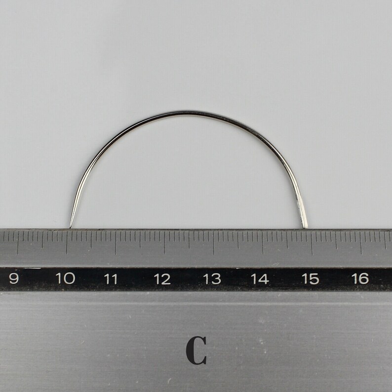 May include: A curved, silver needle measuring 15 inches long. The needle is placed on top of a ruler with markings from 9 to 16.