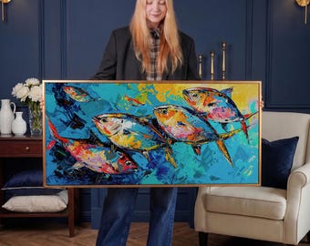 Puede incluir: Pintura enmarcada que representa un grupo de peces coloridos nadando en un mar turquesa. La obra de arte utiliza pinceladas audaces y tonos vibrantes de azul, naranja, amarillo y rojo. La pintura es sostenida por una persona.