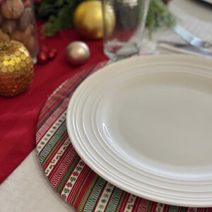 May include: A white dinner plate with a red and green Christmas themed placemat. The placemat has a striped pattern with snowflakes and candy canes.