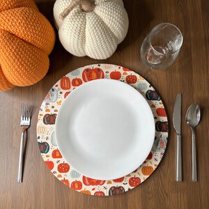 May include: A white and orange pumpkin-themed placemat with a white plate, fork, knife, and spoon on top. The placemat has a cream background with orange, white, and brown pumpkins and leaves.