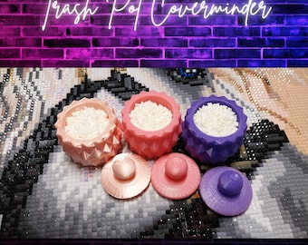 May include: Three colorful Trash Pot Coverminders in pink, coral, and purple, each filled with white beads. Matching lids are in front. The background is a diamond painting with a brick wall and neon sign that reads "Trash Pot Coverminder".