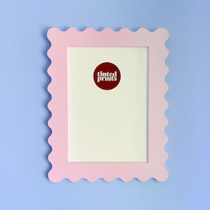 May include: A light pink picture frame with a scalloped edge, mounted on a light blue wall. The frame has a white interior and a circular red sticker with the words "tinted prints".