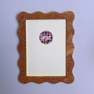 Può includere: Una cornice dorata con glitter e bordo ondulato. La cornice circonda uno spazio bianco con un adesivo circolare al centro che recita "tinted prints" in testo bianco. L'adesivo ha un motivo a rombi rossi, bianchi e blu.
