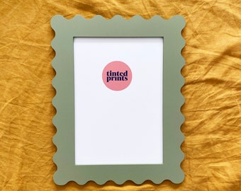 May include: A sage green picture frame with a scalloped edge. The frame is sitting on a yellow fabric surface. A pink circle with the text "tinted prints" is inside the frame.