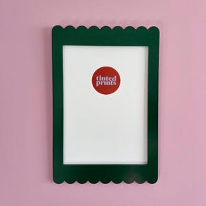 Könnte beinhalten: Ein rechteckiger grüner Bilderrahmen mit gewelltem Rand, montiert an einer rosa Wand. Der Rahmen hat einen weißen Innenraum mit einem roten runden Aufkleber in der Mitte mit der Aufschrift "tinted prints".