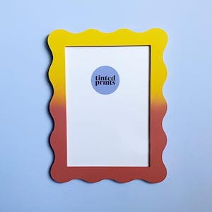 Può includere: Una cornice per foto con un effetto sfumato dal giallo al rosso e un bordo a forma di onda. La cornice ha un passepartout bianco e un piccolo cerchio blu con il testo "tinted prints" al centro.