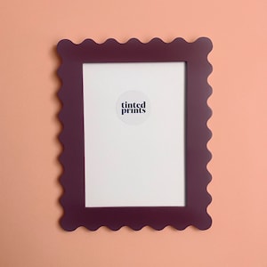 Può includere: Una cornice color prugna con bordo smerlato. La cornice ha un interno bianco con la scritta "tinted prints" su un adesivo circolare. La cornice è esposta su una parete color pesca.
