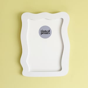 May include: A white, wavy-edged picture frame with a white interior and a circular sticker that reads "tinted prints." The frame is mounted on a pale yellow wall. The frame's design is modern and minimalist.