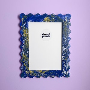 Può includere: Una cornice per foto blu e oro con un bordo a festoni. La cornice ha un adesivo bianco con il testo "Tinted Prints" su di essa.