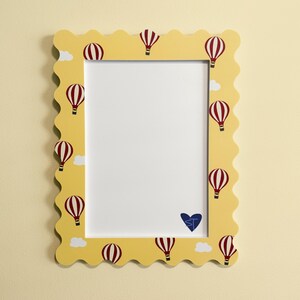 May include: A rectangular picture frame with a scalloped edge, featuring a yellow background decorated with red and white striped hot air balloons and small white clouds. A blue heart with the letters ST is visible in the bottom right corner.