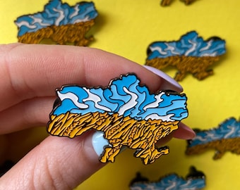 Puede incluir: Un pin de esmalte metálico con la forma del mapa de Ucrania, que presenta un diseño estilizado de cielo azul con nubes blancas sobre un campo de trigo amarillo dorado. El pin tiene un contorno de metal oscuro sobre un fondo amarillo.
