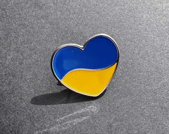 May include: A heart-shaped enamel pin featuring a horizontal split design with a vibrant blue top half and a bright yellow bottom half, enclosed by a thin silver-toned metal border that outlines the entire heart shape.