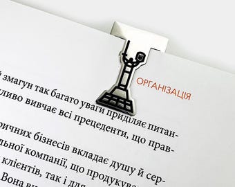 Może przedstawiać: Srebrzysta metalowa zakładka do książki w kształcie pomnika Matki Ojczyzny w Kijowie, przedstawiająca postać trzymającą miecz i tarczę, przypięta do górnej krawędzi otwartej książki z wydrukowanym ukraińskim tekstem na stronie.