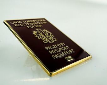May include: A rectangular metal pin featuring a dark burgundy enamel surface with gold-colored trim. The design replicates a Polish passport cover, displaying the text UNIA EUROPEJSKA RZECZPOSPOLITA POLSKA, a central eagle emblem, and the word PASZPORT.