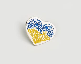 May include: A heart-shaped enamel pin featuring a white background with a floral design. The design consists of blue cornflowers at the top and yellow flowers with green stems at the bottom, all outlined in a metallic silver finish.
