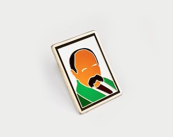 Puede incluir: Un pin de esmalte rectangular con una ilustración estilizada de una persona calva, con rostro naranja y un bigote negro prominente. La figura viste una chaqueta verde y una corbata marrón oscuro sobre un fondo blanco con un borde negro.