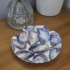 Op de afbeelding: Een decoratieve, schelpvormige schaal met een aquarelontwerp van oesterschelpen in blauwe en paarse tinten. De schaal staat op een houten oppervlak, met een heldere glazen vaas en een witte vaas op de achtergrond.