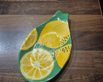 Könnte beinhalten: Ein Keramik-Löffelhalter mit grünem Rand und gelben Zitronenscheiben. Der Löffelhalter ist etwa 20 cm lang und 13 cm breit. Die Zitronenscheiben haben weiße Details und schwarze Kerne.