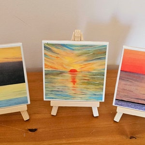 Könnte beinhalten: Drei kleine Gemälde auf Staffeleien, die Meerblicke mit Sonnenuntergängen darstellen. Die Gemälde sind auf quadratischen Leinwänden mit weißem Hintergrund. Das erste Gemälde zeigt einen dunkelblauen Himmel mit einem gelben Sonnenuntergang und einem hellblauen Meer. Das zweite Gemälde zeigt einen leuchtend orangenen Sonnenuntergang mit einem hellblauen Meer. Das dritte Gemälde zeigt einen roten Sonnenuntergang mit einem hellblauen Meer.