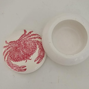Op de afbeelding: Een witte, ronde sieradendoos met een deksel met een rode krabillustratie. De doos is open en toont een cirkelvormig interieur. Het krabontwerp is gedetailleerd en bedekt het grootste deel van het oppervlak van het deksel.