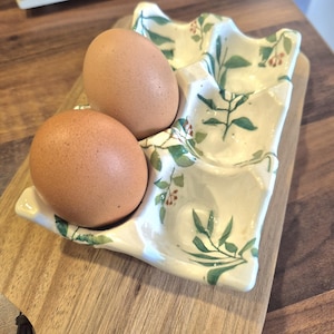 Może przedstawiać: Dwa brązowe jajka spoczywają w białym ceramicznym pojemniku na jajka z kwiatowym wzorem. Pojemnik znajduje się na drewnianej desce do krojenia. Pojemnik na jajka ma zakrzywiony kształt z trzema przegródkami i jest ozdobiony zielonymi liśćmi i czerwonymi jagodami.