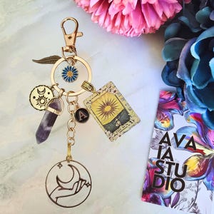 May include: A gold-toned keychain featuring a lobster clasp, a blue and yellow flower charm, a gold feather, a round sun and moon charm, a purple crystal point, a small black letter A charm, and a rectangular tarot card charm labeled The Sun.