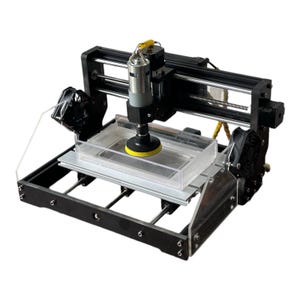 May include: A black and silver CNC machine with a clear plastic work area and a yellow sanding pad attached to the spindle.
