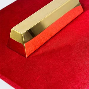 Può includere: Una barra in metallo color oro lucido con sezione trasversale trapezoidale e superficie liscia e riflettente. La barra è posizionata diagonalmente su una superficie rosso vivo dalla consistenza vellutata, che ne esalta la lucentezza.