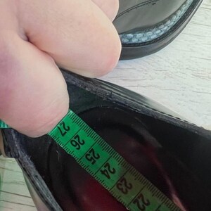 May include: A black shoe with a smooth finish and a textured welt detail. A green measuring tape is placed inside the shoe, showing a length measurement of approximately 27 centimeters from the heel to the toe area of the interior footbed.