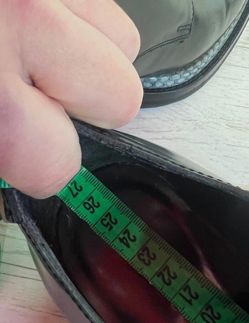 May include: A black shoe with a smooth finish and a textured welt detail. A green measuring tape is placed inside the shoe, showing a length measurement of approximately 27 centimeters from the heel to the toe area of the interior footbed.