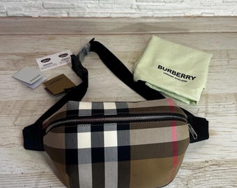 May include: A beige, black, and white plaid fanny pack with a black strap and zipper. The bag has a curved shape and a silver zipper. A beige cloth bag with "BURBERRY LONDON ENGLAND" printed on it is also present.