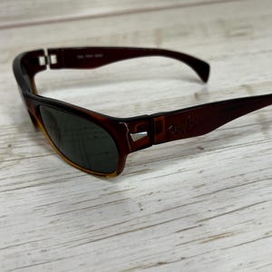 May include: Brown and black sunglasses with a tortoiseshell pattern. The sunglasses have dark lenses and the Ray-Ban logo on the right arm. The frames are a dark brown color.