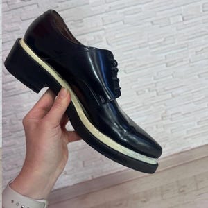 May include: A black lace-up shoe with a glossy finish, featuring a thick, layered sole with a distinct white middle section and a black bottom tread. The shoe has a rounded toe, a low-cut ankle opening, and black laces secured through eyelets.