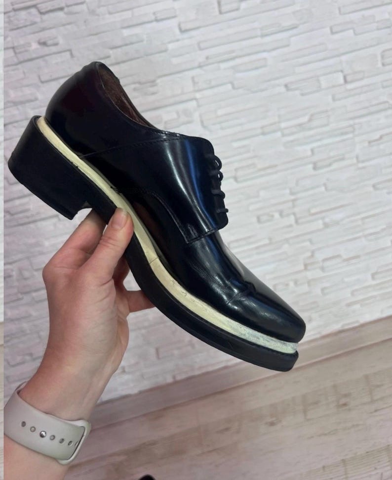 May include: A black lace-up shoe with a glossy finish, featuring a thick, layered sole with a distinct white middle section and a black bottom tread. The shoe has a rounded toe, a low-cut ankle opening, and black laces secured through eyelets.