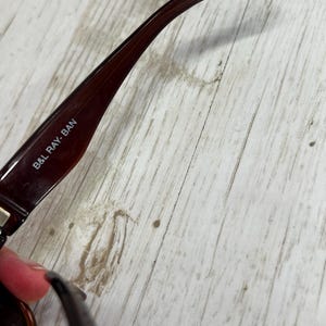 May include: Close-up of a pair of brown sunglasses with the text "B&L RAY-BAN" printed on the arm. The sunglasses have a dark brown frame and a curved arm. The background is a light-colored wooden surface.