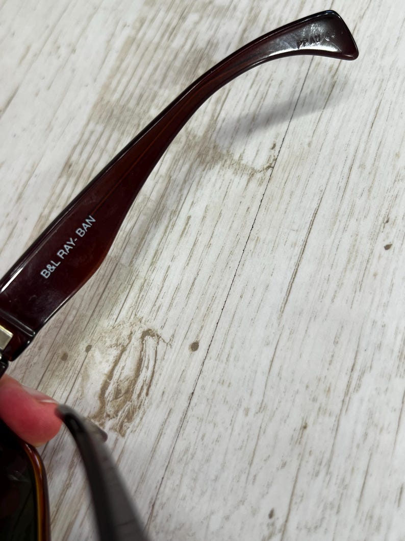 May include: Close-up of a pair of brown sunglasses with the text "B&L RAY-BAN" printed on the arm. The sunglasses have a dark brown frame and a curved arm. The background is a light-colored wooden surface.